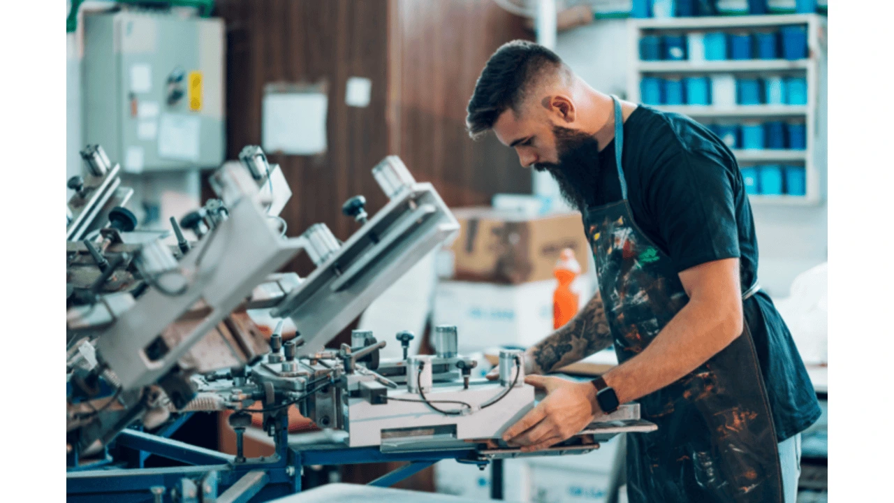 A person working at a printing press, focusing on a machine used for screen printing, with materials and tools visible in the background